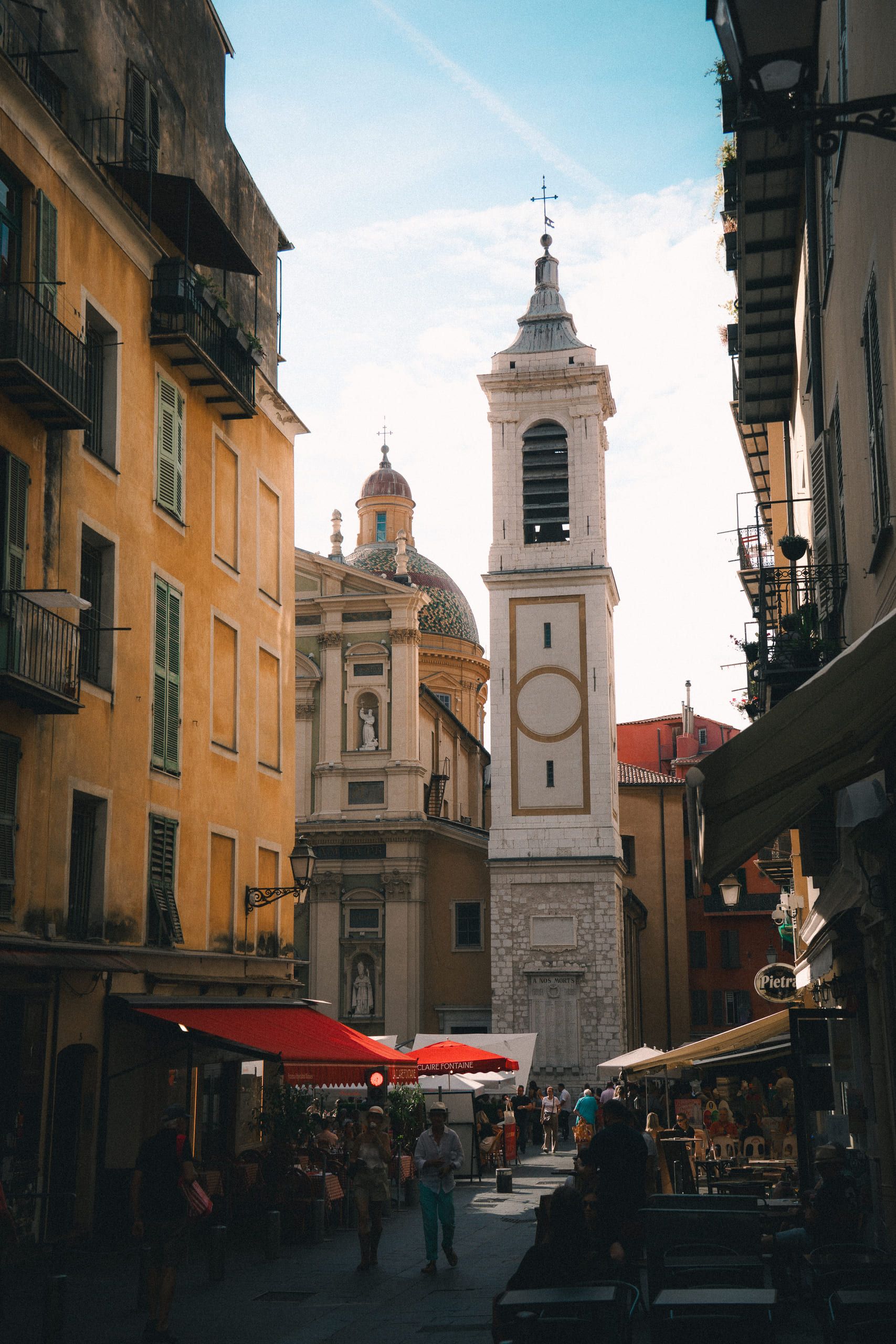A bell tower and a tiled baroque dome close the end of a narrow Old Town street in Nice, café awnings and terrace tables below.