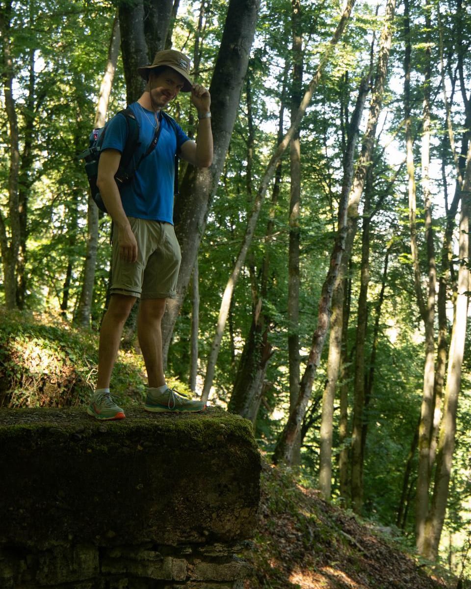Yaroslav standing on a mossy stone ledge in a sunlit beech forest, tipping the brim of his hat towards the camera.
